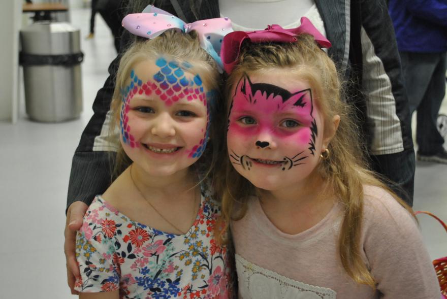 Two children with face paint smiling brightly, one with a fish design and the other with a cat.
