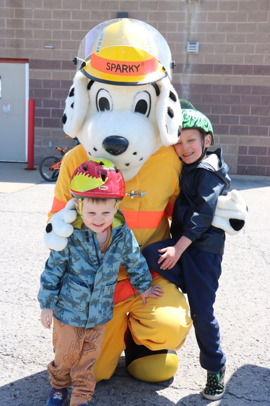 Children posing with a costumed mascot wearing a helmet, outdoors on a sunny day.