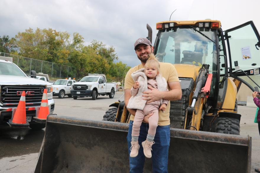 A person holds a child in front of a construction vehicle with trees and clouds in the background.