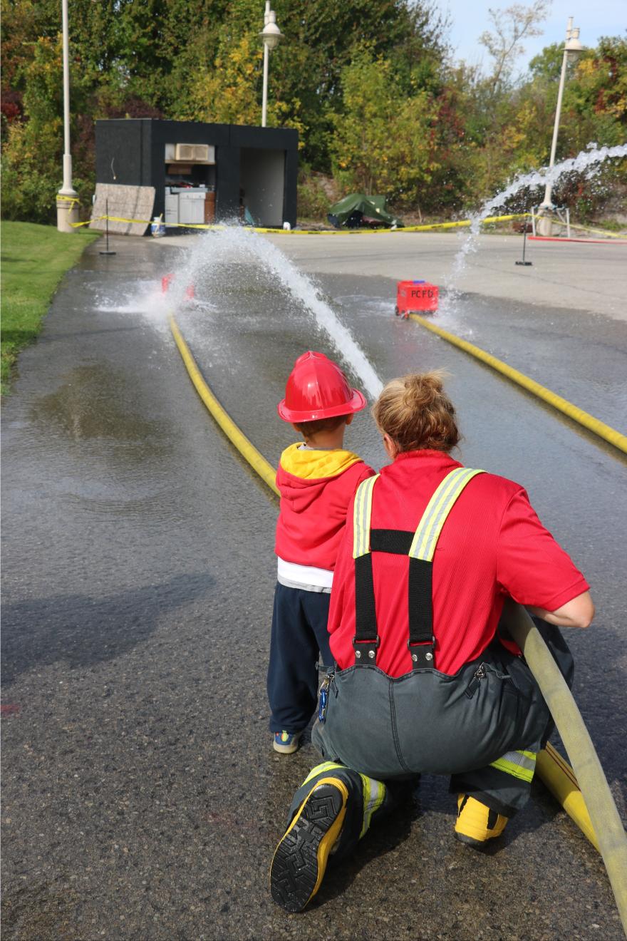 A person and a child in a red helmet kneel beside a fire hose, spraying water in a park.
