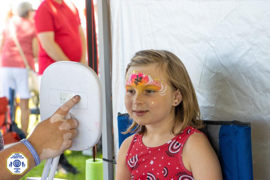 A child sits in a chair, smiling while receiving face painting at an outdoor event.