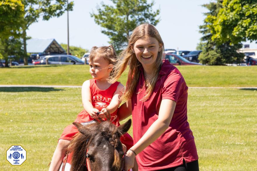 A person leads a pony while a child rides, smiling in a sunny park.