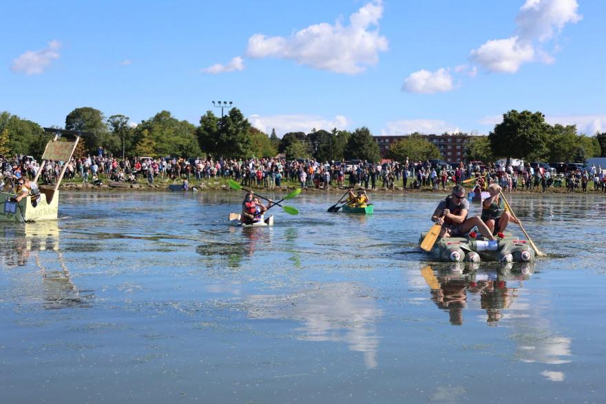 Participants in boats paddle across a calm waterway, surrounded by a large crowd on shore.