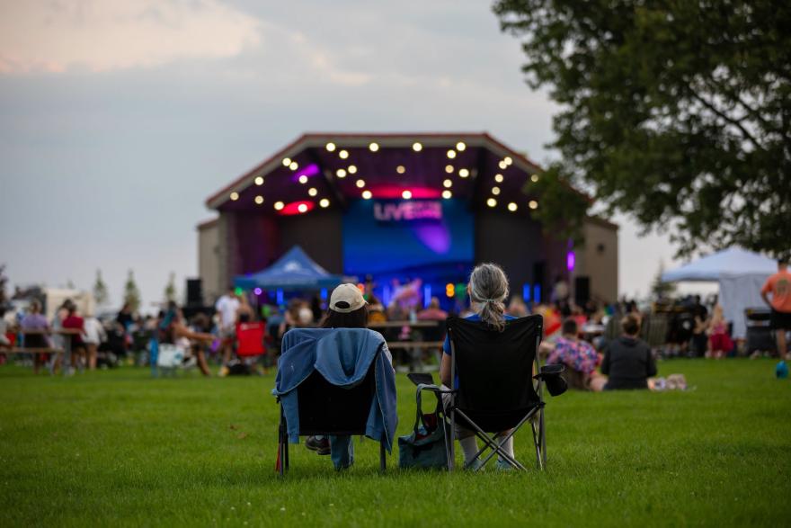 Two people sit on folding chairs, enjoying a live outdoor concert on a grassy field.