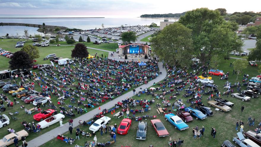 Aerial view of a large outdoor concert with a crowd sitting on grass and classic cars parked nearby.