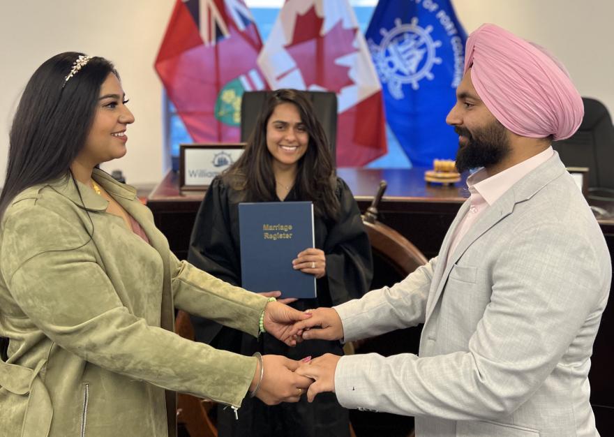 Couple holding hands during a ceremony, with a judge in the background and flags displayed.