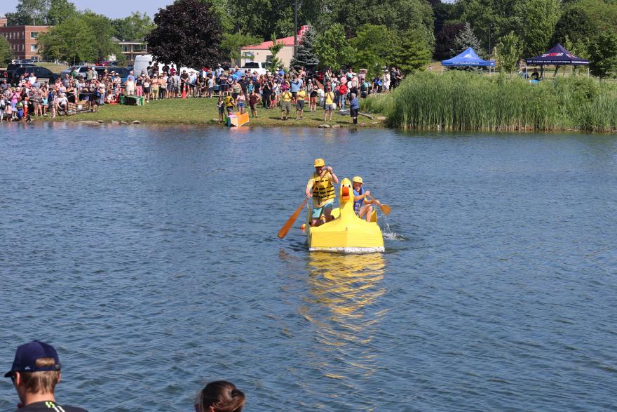 Participants in a yellow boat paddling across a lake, with a crowd watching onshore.