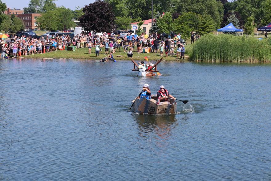 Participants paddle homemade rafts while spectators watch from the grassy shoreline.