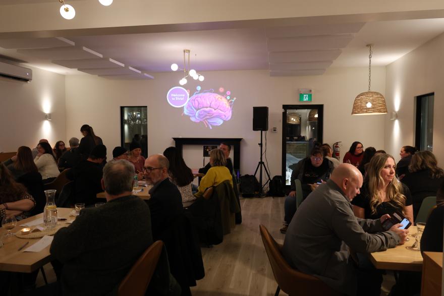 A lively indoor gathering with people seated at tables and a projection on the wall.