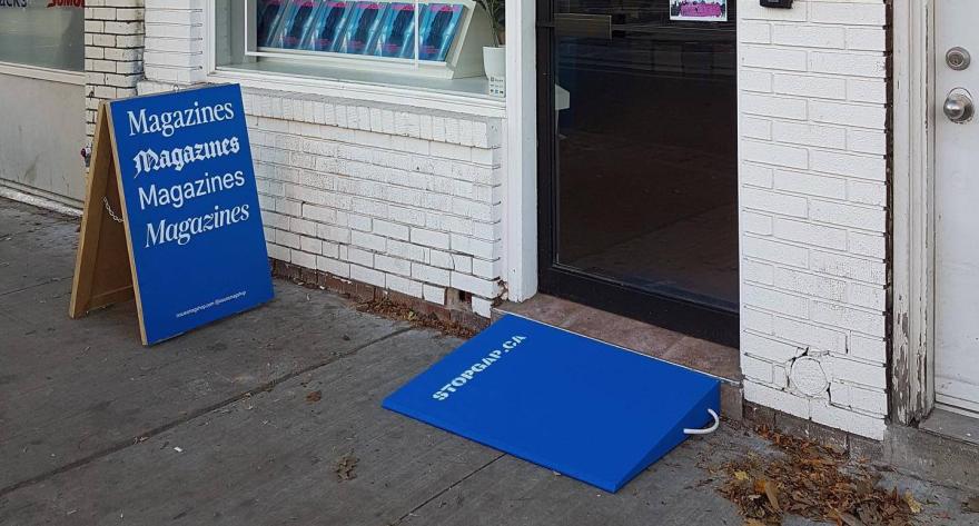 Blue ramp and sign outside a magazine store, with autumn leaves on the sidewalk.