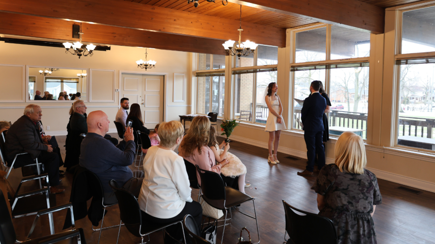 A couple stands together in front of a small audience in a warmly lit venue.