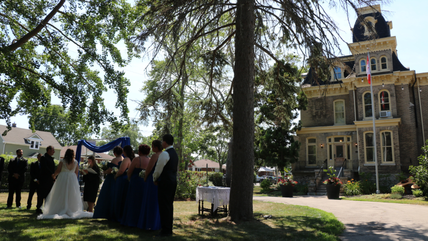 Outdoor wedding ceremony in front of an ornate building, surrounded by trees and greenery.