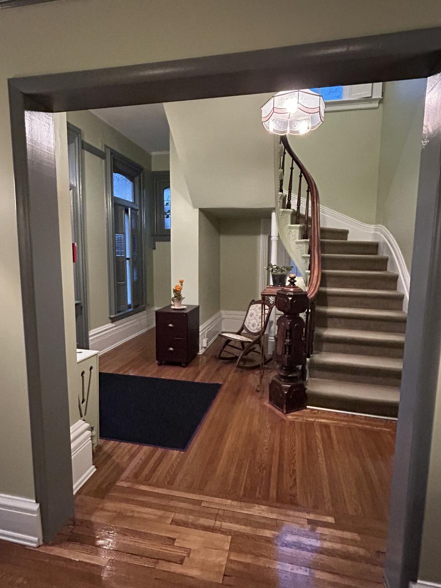 A view through an arched doorway into a well-lit hallway with stairs, hardwood floors, and a small table.