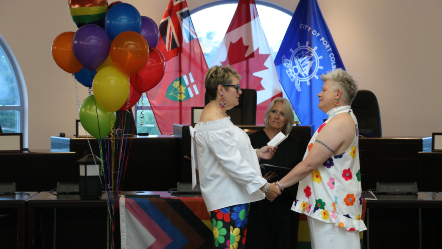 Two people hold hands during a wedding ceremony, with colourful balloons and flags in the background.