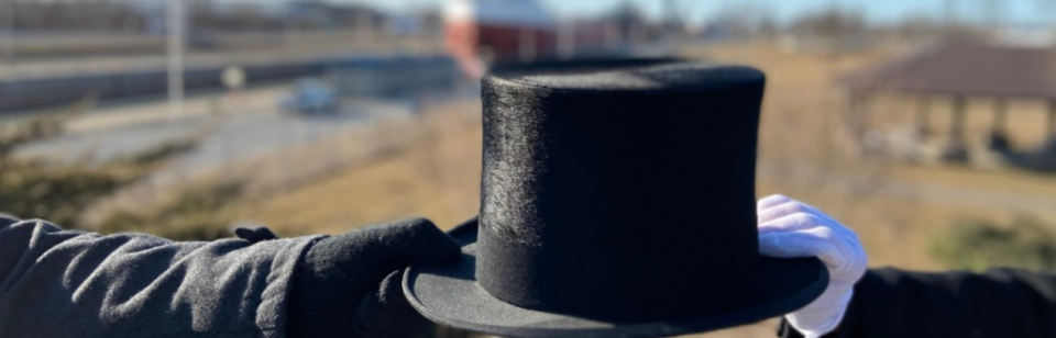 A top hat is held between two gloved hands, with a park and road in the background.