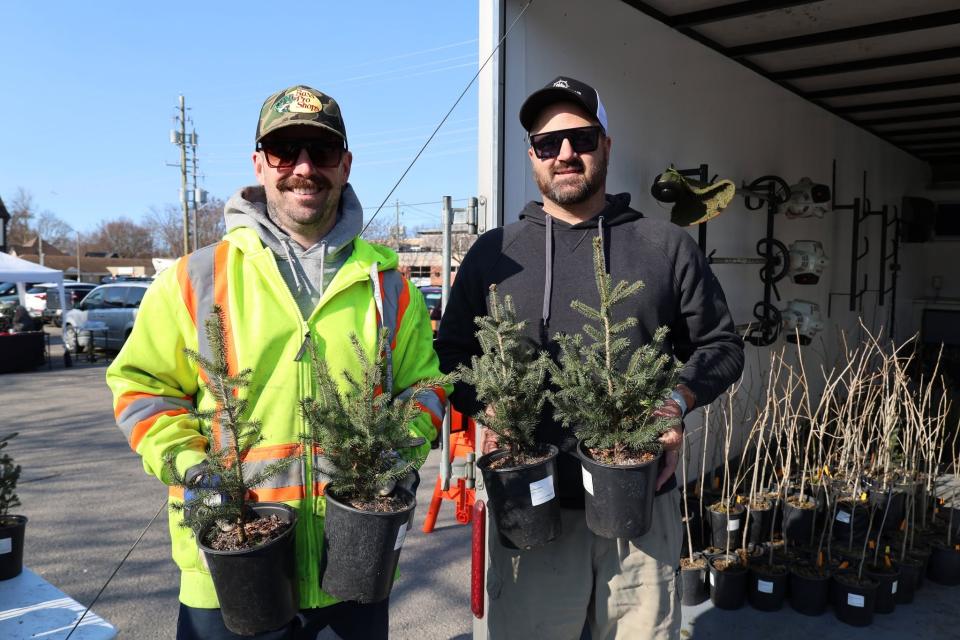 Two individuals hold potted trees, standing outdoors beside a trailer and work equipment.