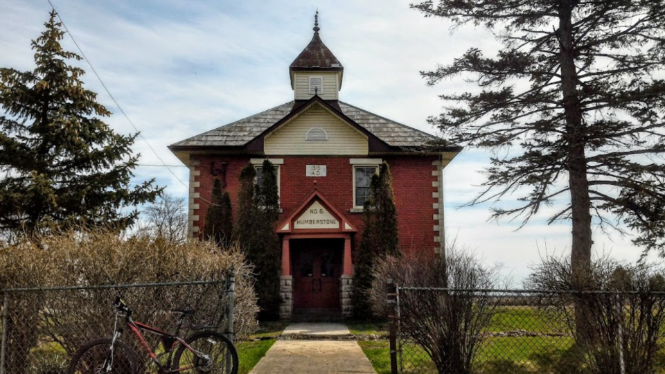Historic red brick building with a pointed roof, surrounded by trees and a bike in front.