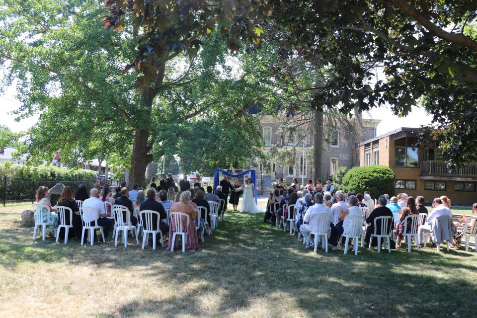 Outdoor wedding ceremony with guests seated on white chairs under trees.