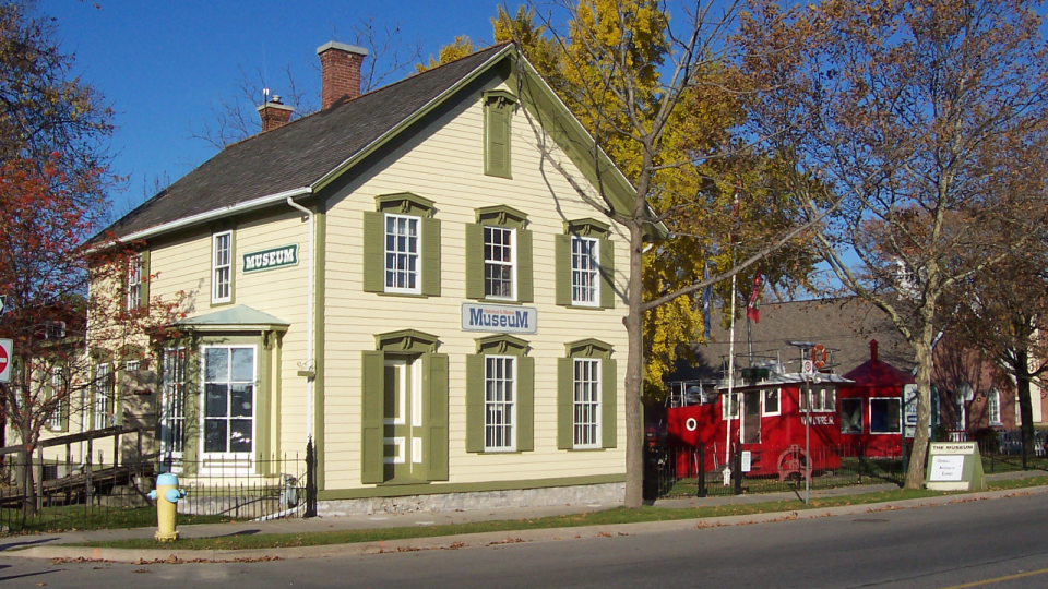 Historic yellow house with green accents, red structure nearby, autumn foliage.