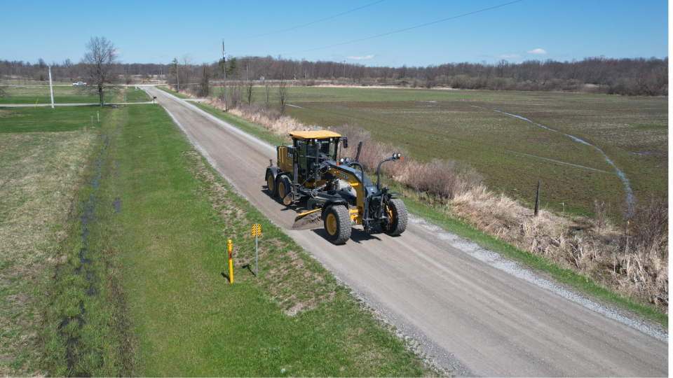 Grader machine working on a gravel road beside a green field on a sunny day.