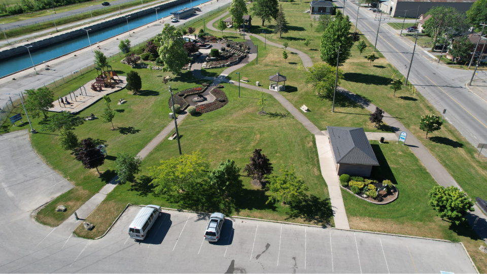 Aerial view of a green park with pathways, trees, and a small pavilion near a canal.