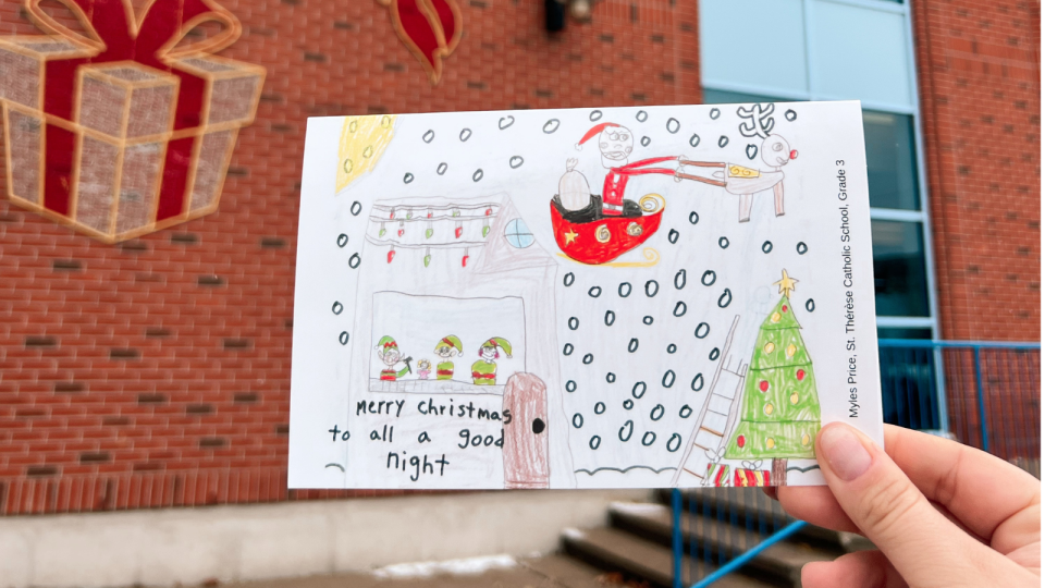 A hand holding a colourful Christmas card with a festive drawing in front of a brick wall.