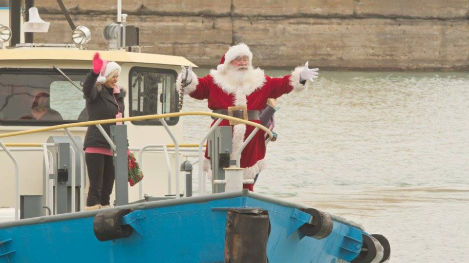 Santa Claus waves from a boat while a person stands beside, both smiling near water.