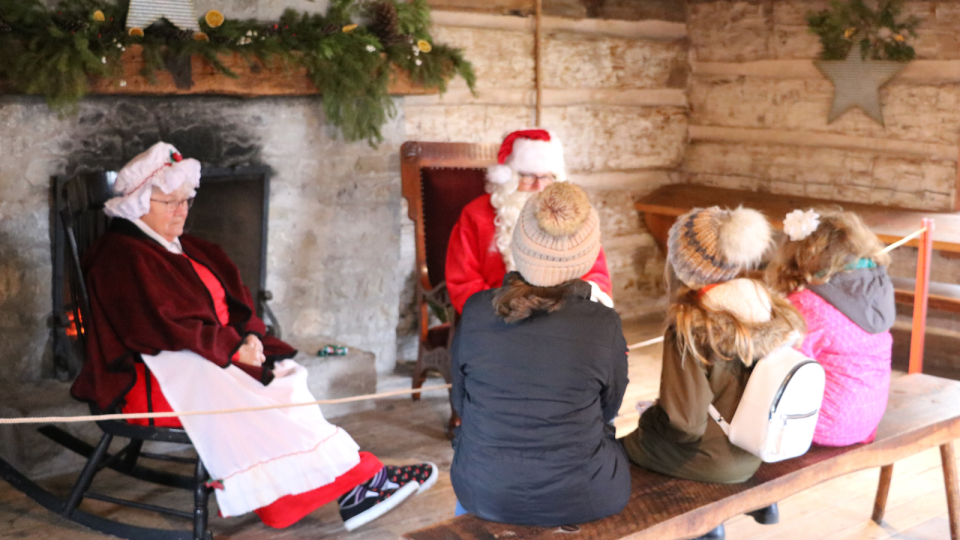 Santa Claus and a companion greet children seated on a bench in a festive cabin.