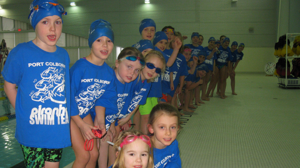 A group of children in blue swim shirts and caps, lined up by a pool.