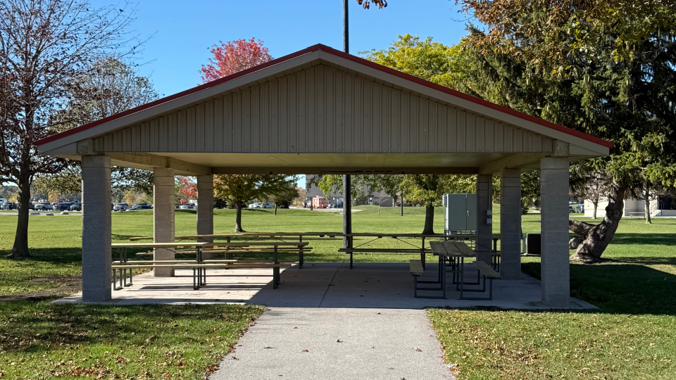 A picnic shelter in a park with benches and tables, surrounded by green grass and autumn trees.