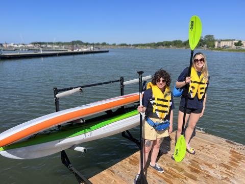 Two individuals wearing life jackets stand on a dock beside a kayak and paddles, with water behind.