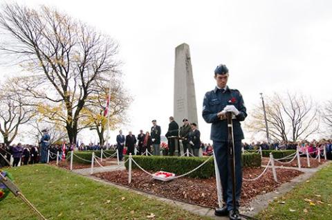 A solemn ceremony at a war memorial with a crowd in the background and trees in autumn.