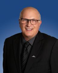 Business professional with glasses, wearing a black suit, smiling against a blue background.