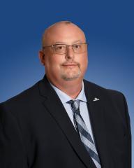 A person wearing glasses and a suit, smiling against a blue background.