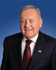 Formal portrait with a smiling individual in a dark suit and a patterned tie against a blue background.