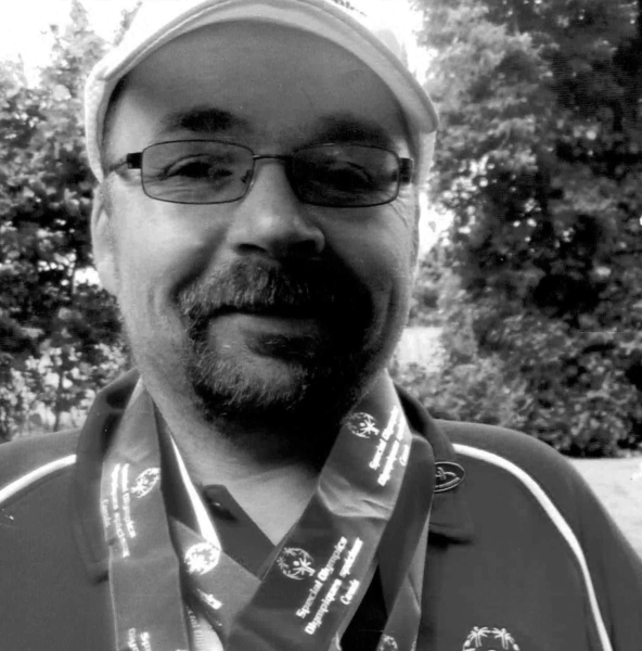 Person smiling, wearing glasses and a cap, with medals around their neck in a leafy background.