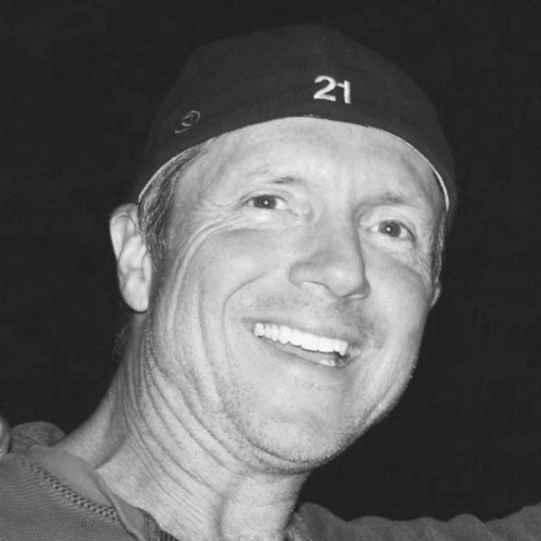 Smiling individual wearing a black cap, against a dark background.