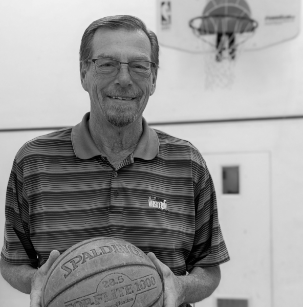 A smiling person holds a basketball in a gym with a hoop visible in the background.