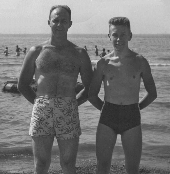 Two people stand by the water in vintage swimwear, with others swimming in the background.