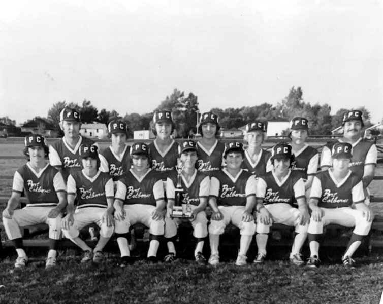 A group of eleven individuals in matching baseball uniforms posing with a trophy on a field.