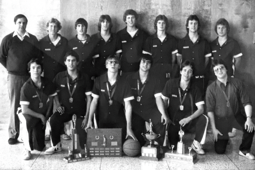 A black-and-white photo of a basketball team with trophies, standing and kneeling together.
