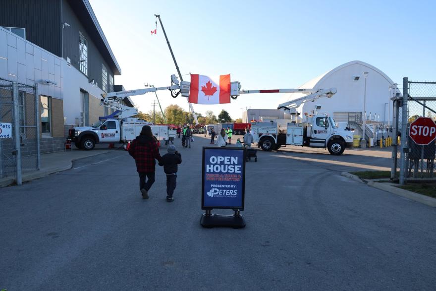 Two people walk towards a sign for an open house, with Canadian flags in the background.