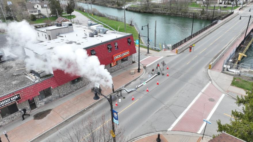 Steam billowing from a building near a road with orange cones and a river in the background.